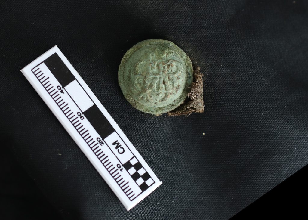 This gilded brass coat button and fabric comes from a Grand Army of the Republic (GAR) uniform and was buried with an African American Civil War veteran who died and was buried in the national cemetery between 1890-1891. This object will be reburied with the remains of the veteran it was found with.