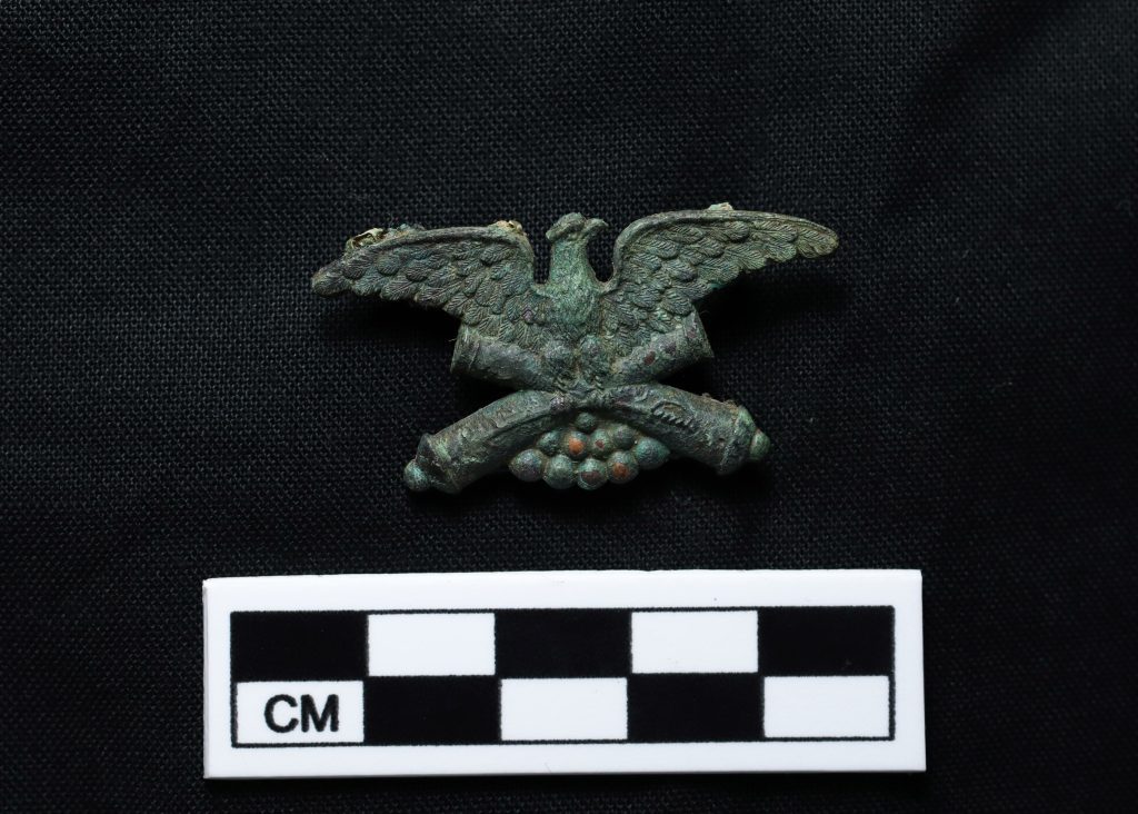 This is the top half of a gilded bronze GAR medal with pin mechanism found still in place on the left lapel of the coat of a USCT veteran who died and was buried in 1890-1891 in the national cemetery. The GAR medal is very similar in its design to the Medal of Honor granted by Congress. This object will be reburied with the remains of the veteran it was found with.